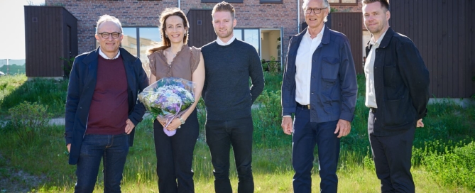 Fem personer står smilende foran et grønt område med nybyggede boliger i baggrunden. En kvinde holder en buket blomster.