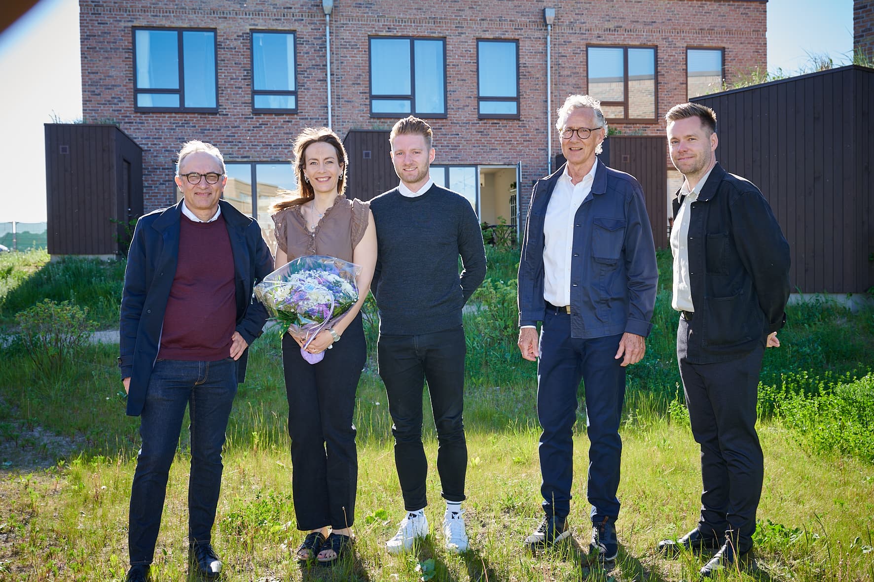 Fem personer står smilende foran et grønt område med nybyggede boliger i baggrunden. En kvinde holder en buket blomster.